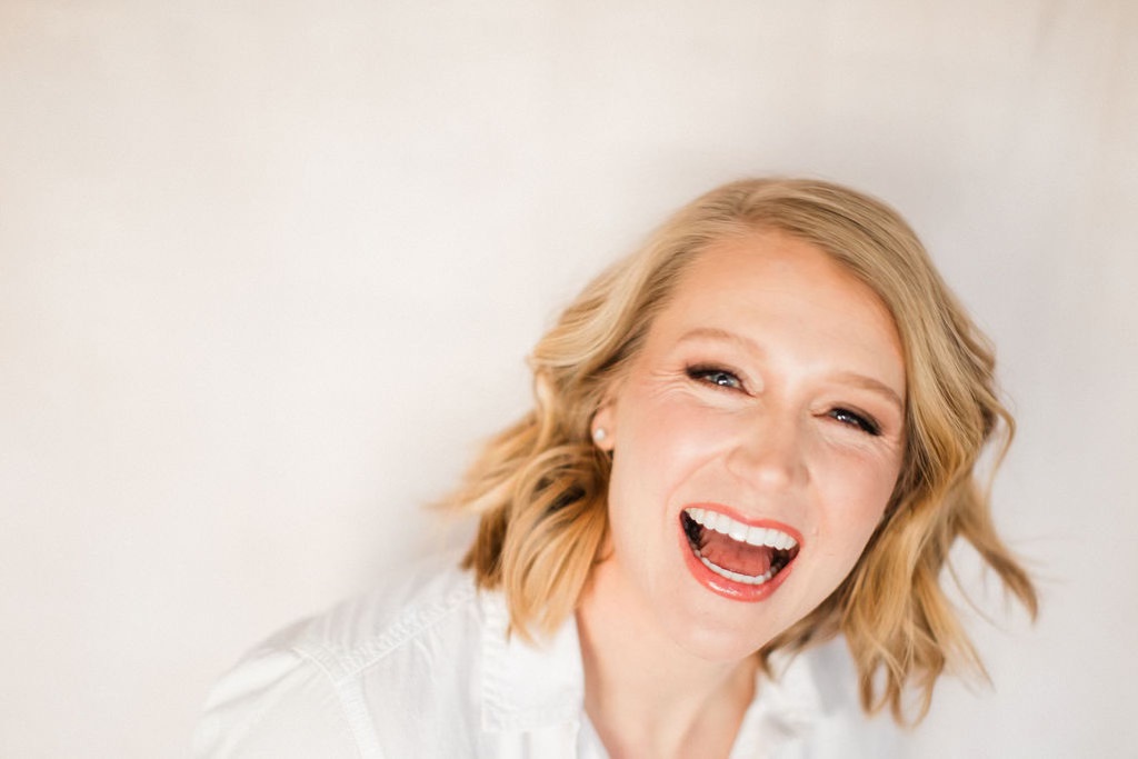 Blonde girl in white shirt with red lipstick laughing with tan background
