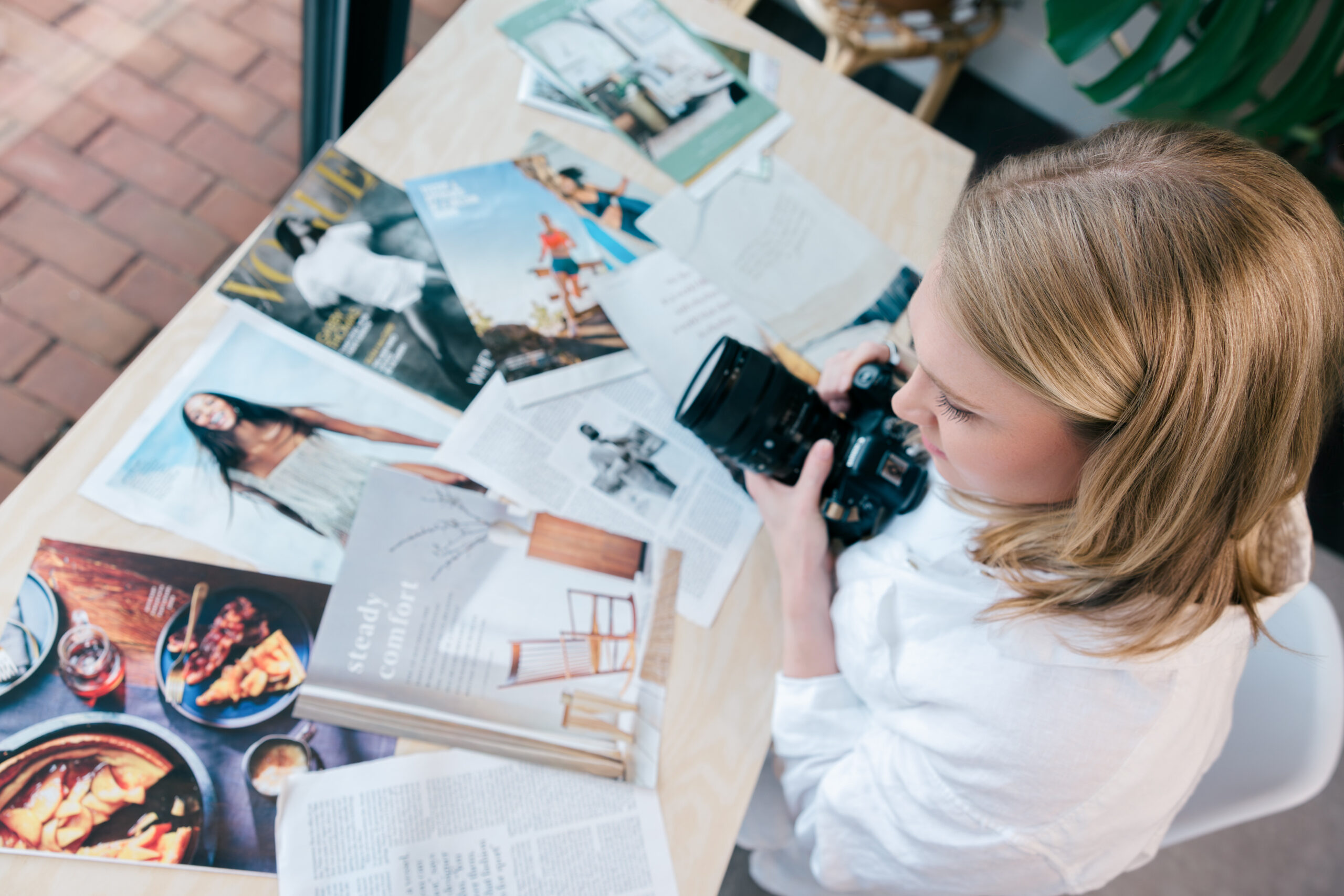blonde girl editor in cheif of digital magazine with her camera looking at magazine clippings on table.
